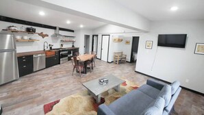 A modern kitchen with wooden cabinets, a dining area with a wooden table and chairs, a gray sofa, and a flat-screen TV mounted on the wall.