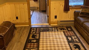 A cozy living room with a large rug featuring animal prints, a couch, and a wooden cabinet. A door leads to a hallway with a sign reading "LAKE HOUSE". - Lake view , Cozy One Bedroom! Walk to Beach and Town! (Bolton)