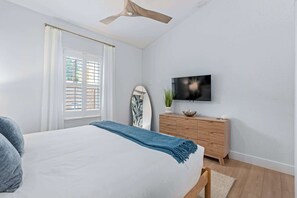 Une chambre à coucher avec un lit, un ventilateur de plafond, une télévision, une commode et une fenêtre avec des rideaux.