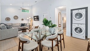A modern living room with a glass dining table, grey sofa, and a washing machine in the background. - Citrus Hideaway l Hot Tub l 1 Block to Downtown (St. Augustine)