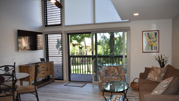 A spacious living room with a glass coffee table, a sofa, a dining table with chairs, and a TV mounted on the wall. - Beautifully remodeled 2 Bd, 2 Bath Condo, short walk to the Beach (Kihei)
