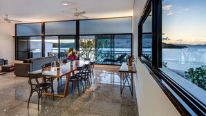 Un espace repas moderne avec une grande table et des chaises, un canapé et une vue sur un plan d’eau grâce à de grandes fenêtres.