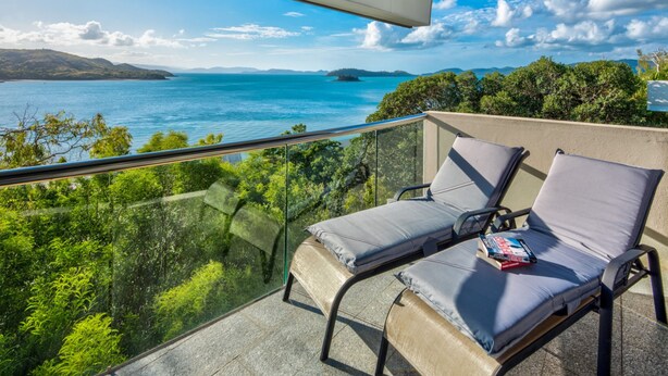 Un balcon avec deux fauteuils confortables et un livre, offrant une vue panoramique sur une étendue d’eau et un paysage lointain.