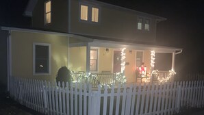 Ein zweistöckiges Haus mit weißem Zaun, einer Veranda mit Lichterketten und einer gut sichtbaren Haustür.