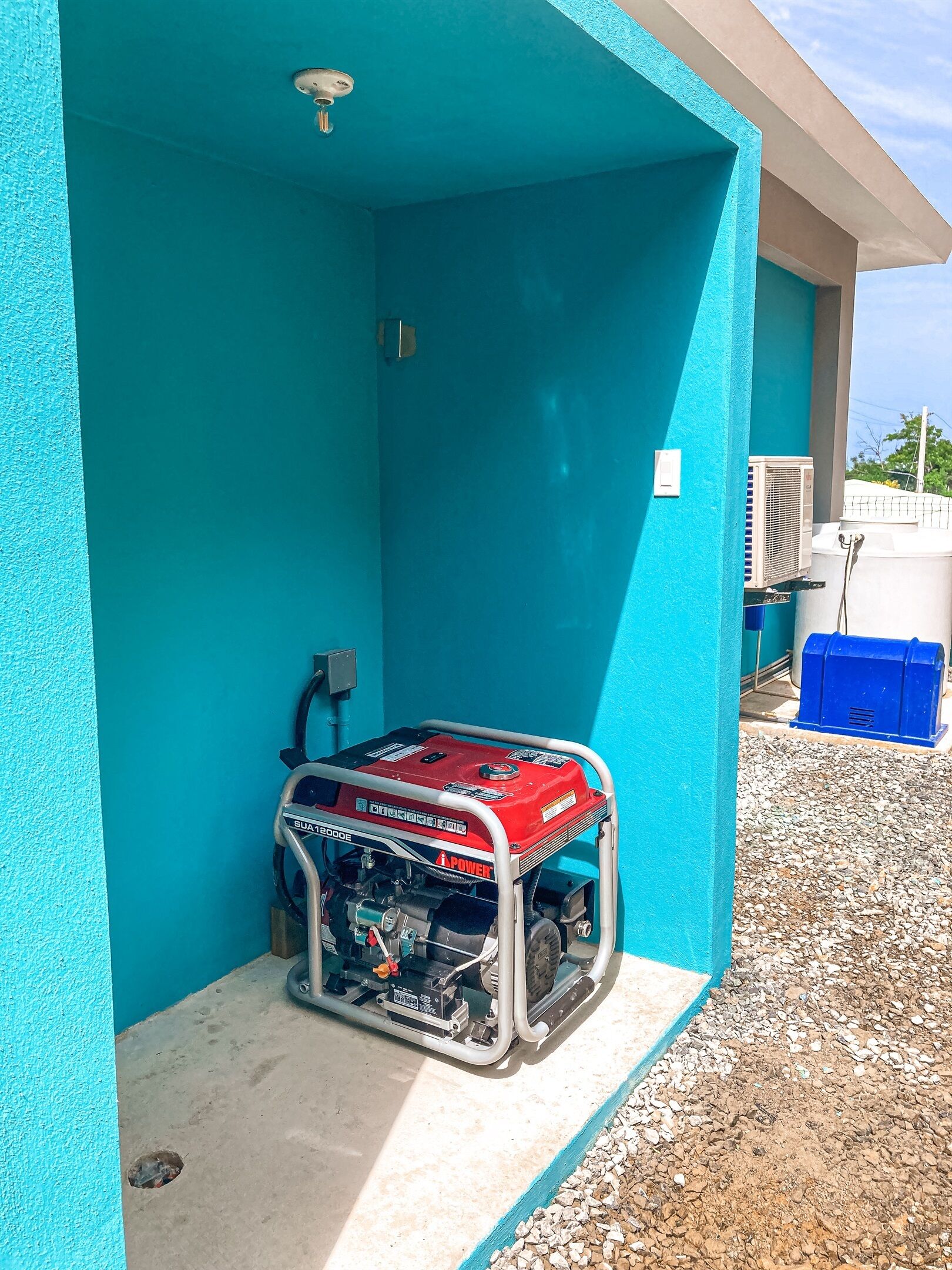 Un generador portátil en un espacio al aire libre techado, con una pared azul, suelo de concreto y un camino de grava.