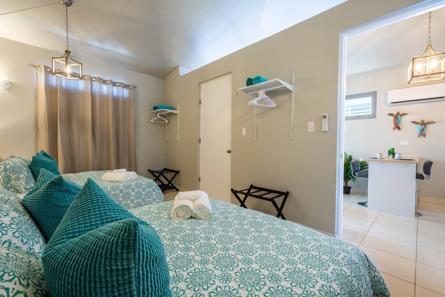 A hotel room with two beds, a desk, a chair, a hanging light fixture, and a door leading to a bathroom.