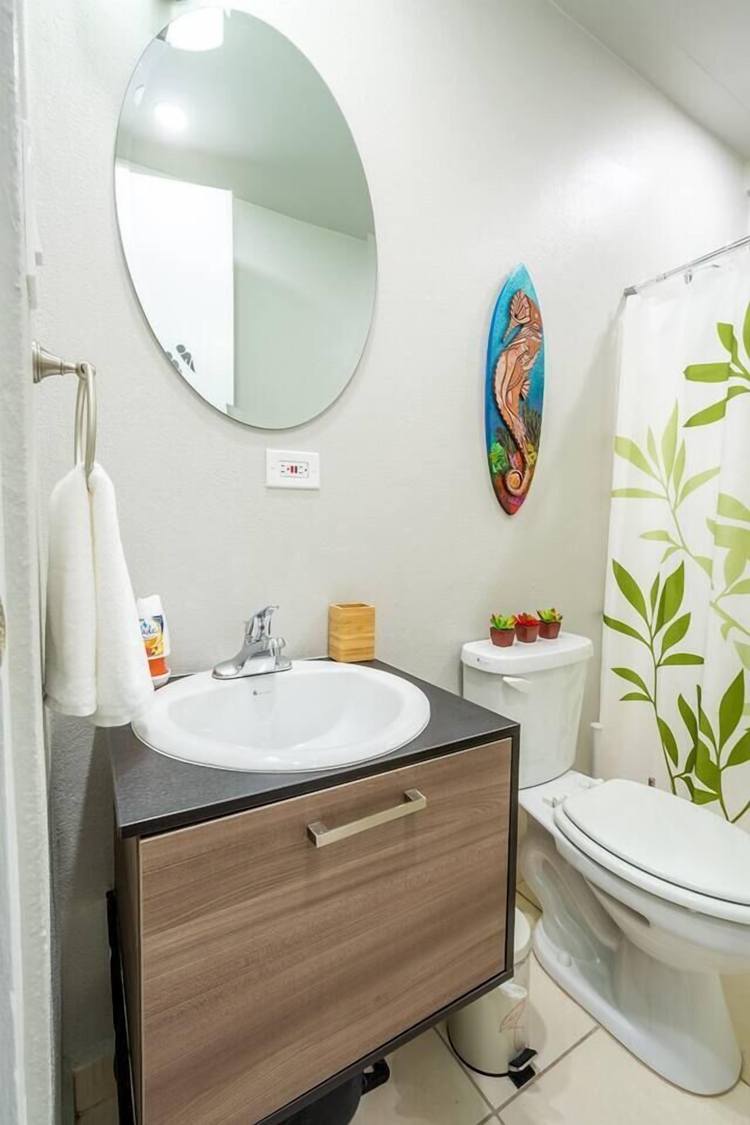 A bathroom with a white sink, a wooden cabinet, a mirror, a toilet, and a shower curtain with a leaf pattern.
