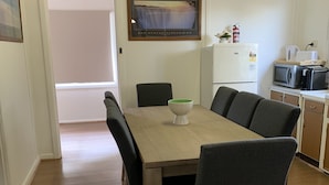 Een eetruimte met een houten tafel en zes stoelen, een lijst met foto aan de muur, en een keuken met apparatuur.