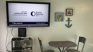 Une pièce avec un téléviseur fixé au mur diffusant une publicité, une petite table avec deux chaises et une étagère contenant divers objets.