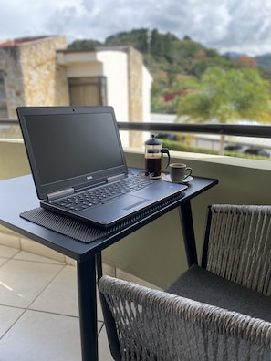 Una laptop sobre una mesa con una cafetera y una taza, con vista a un balcón que da a edificios y árboles.