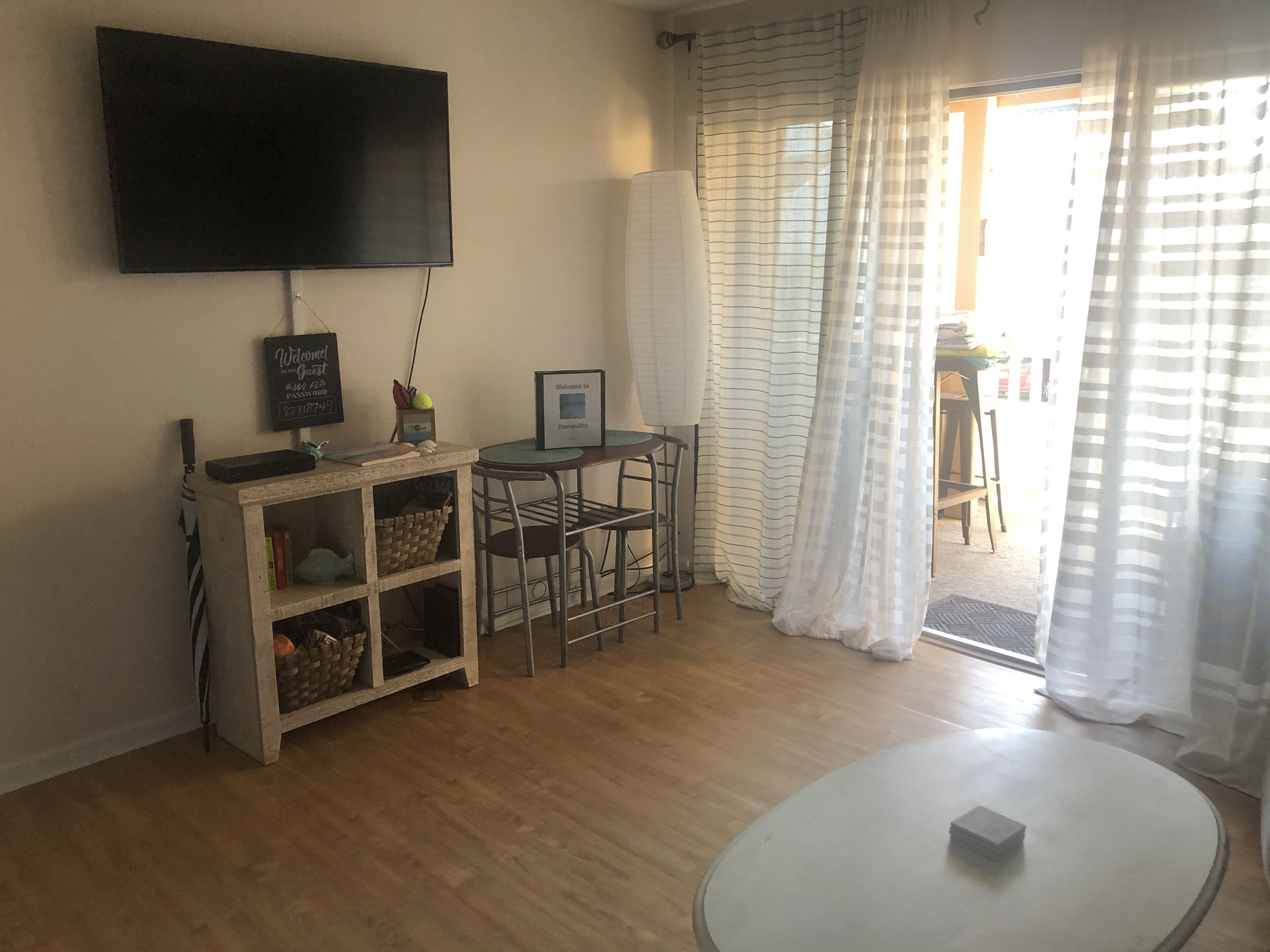A living room with a flat-screen TV, a small table, a chair, a bookshelf, and a window with curtains.