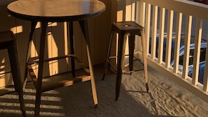 A wooden balcony with a round table and two stools, overlooking a residential street with houses and parked cars.
