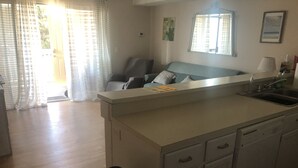 A kitchen with a central island, a sink, and a trash bin. There is a chair and a window with curtains.