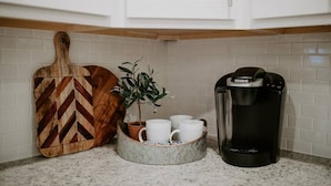 A kitchen counter with a wooden cutting board, a potted plant, a coffee maker, and a marble tray holding cups.