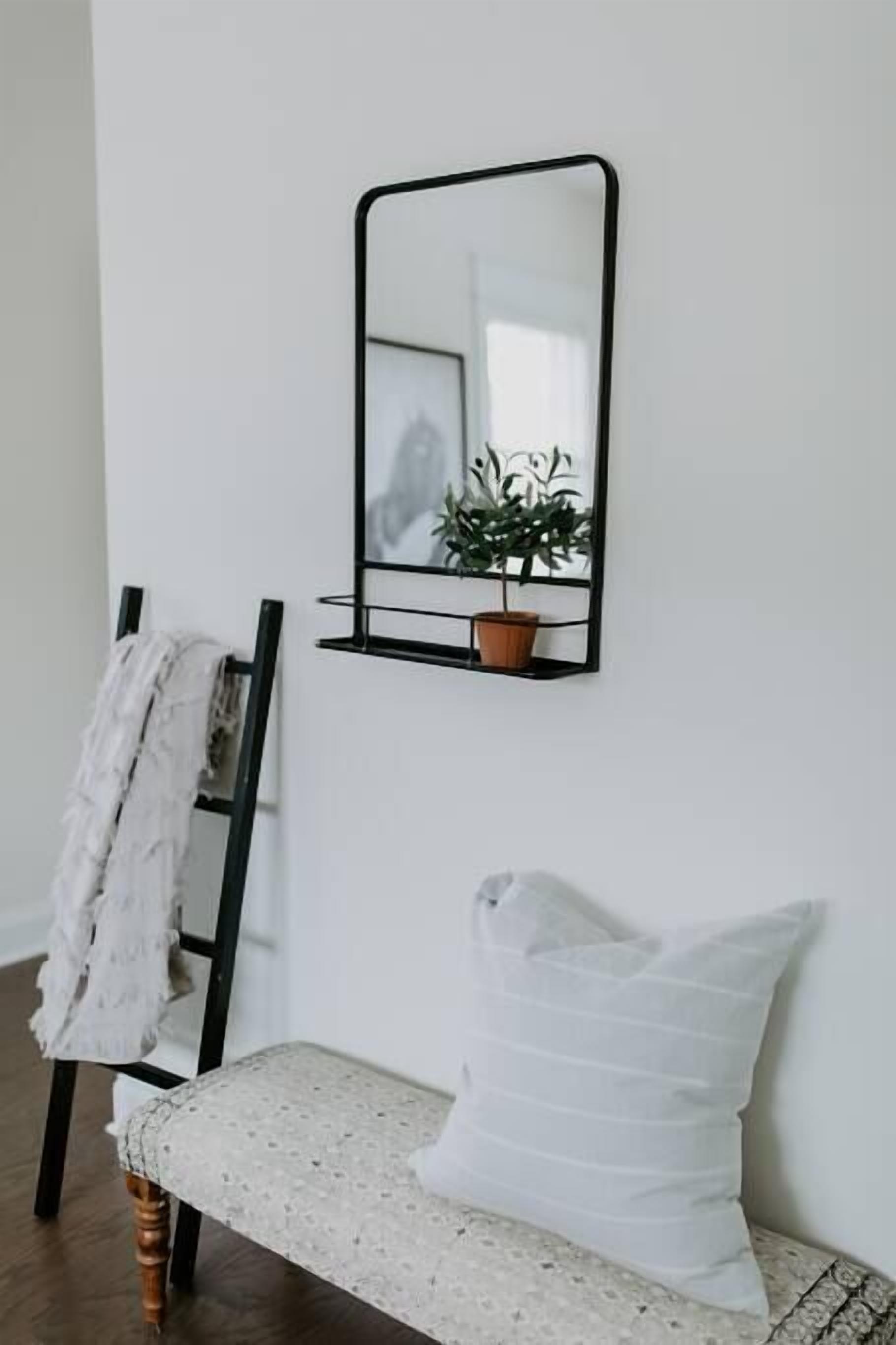 A white wall with a black-framed mirror, a potted plant, a bench with a white cushion, and a wooden chair with a white cloth.