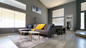 A spacious living room with a grey sofa, a coffee table, and a rug. There are large windows with blinds, a floor lamp, and a painting on the wall. - Perfect Vegas Oasis with Gorgeous Yard/Spa and Pool, Remote Working & Vacation (Henderson)