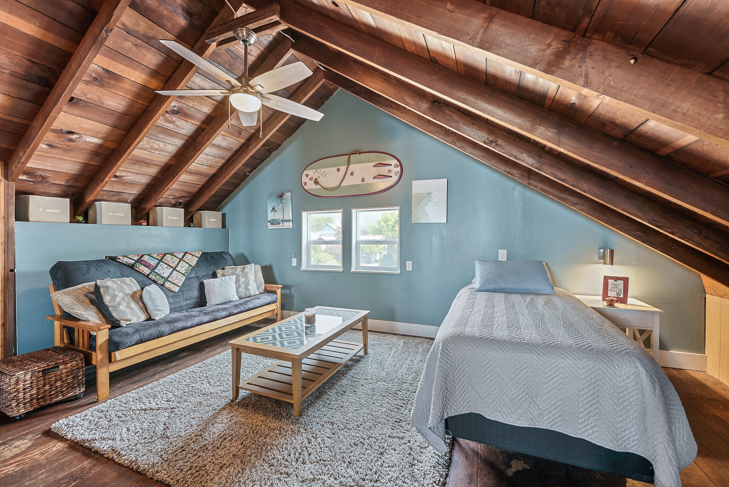 An attic bedroom with a sloped ceiling, a wooden ceiling fan, a sofa, a bed, a small table, and a window.