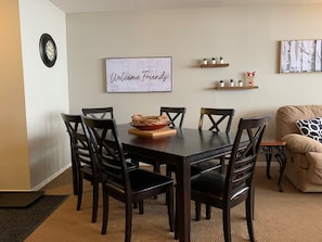 Un comedor con una mesa de madera oscura, seis sillas, un letrero de bienvenida y una pequeña estantería con plantas.