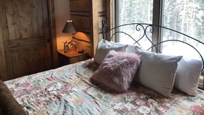 Une chambre avec une porte en bois, une lampe sur une table de chevet, un lit avec une literie à motifs floraux et une tête de lit en métal, et une fenêtre donnant sur une forêt.
