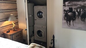 Une pièce comprenant un lit, une table de chevet en bois avec une lampe, un tableau représentant des chevaux accroché au mur, et un espace buanderie avec deux machines à laver.