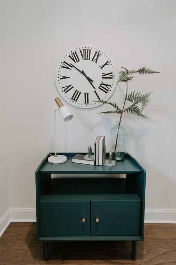 A teal sideboard with a large wall clock, a floor lamp, and decorative items including books and a plant.
