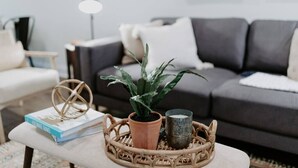 Una sala de estar con un sofá gris oscuro, un sillón blanco y una mesita con un libro, una planta en maceta y una vela.