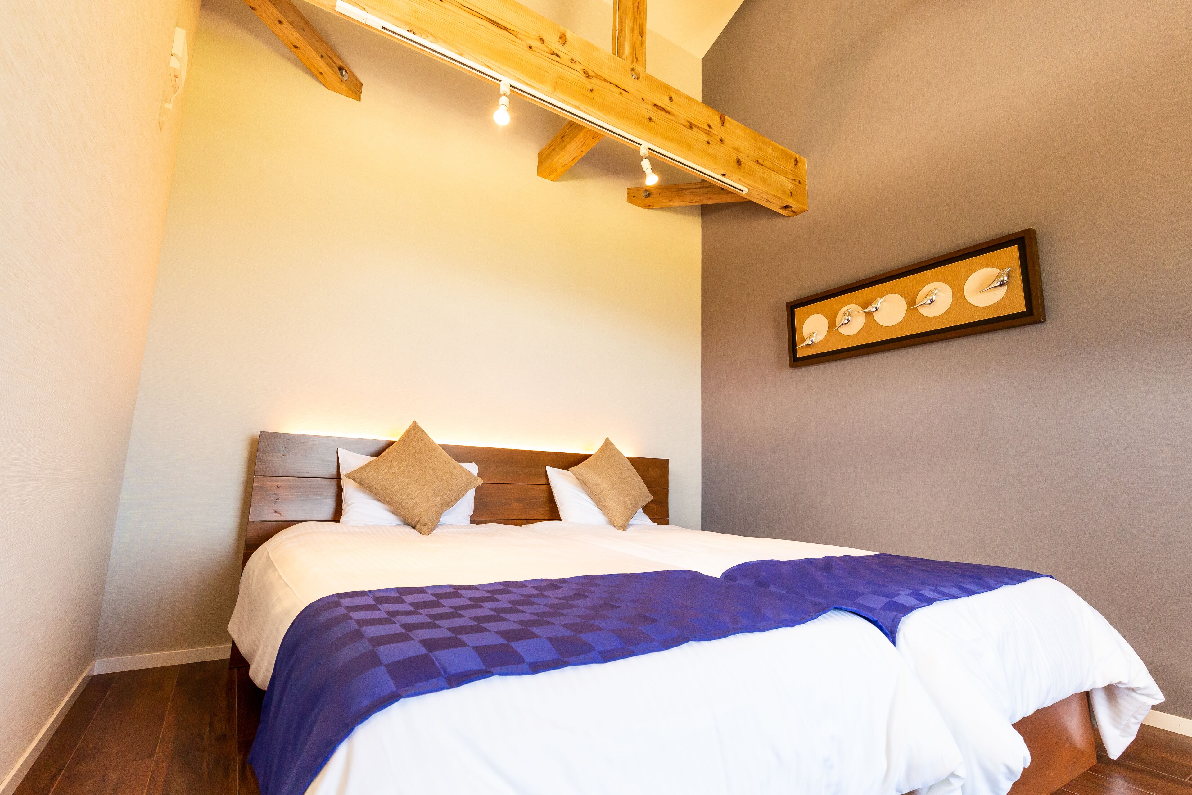 A neatly made bed with a blue checkered blanket and two pillows, a wooden headboard, and a wall-mounted picture of circular objects.