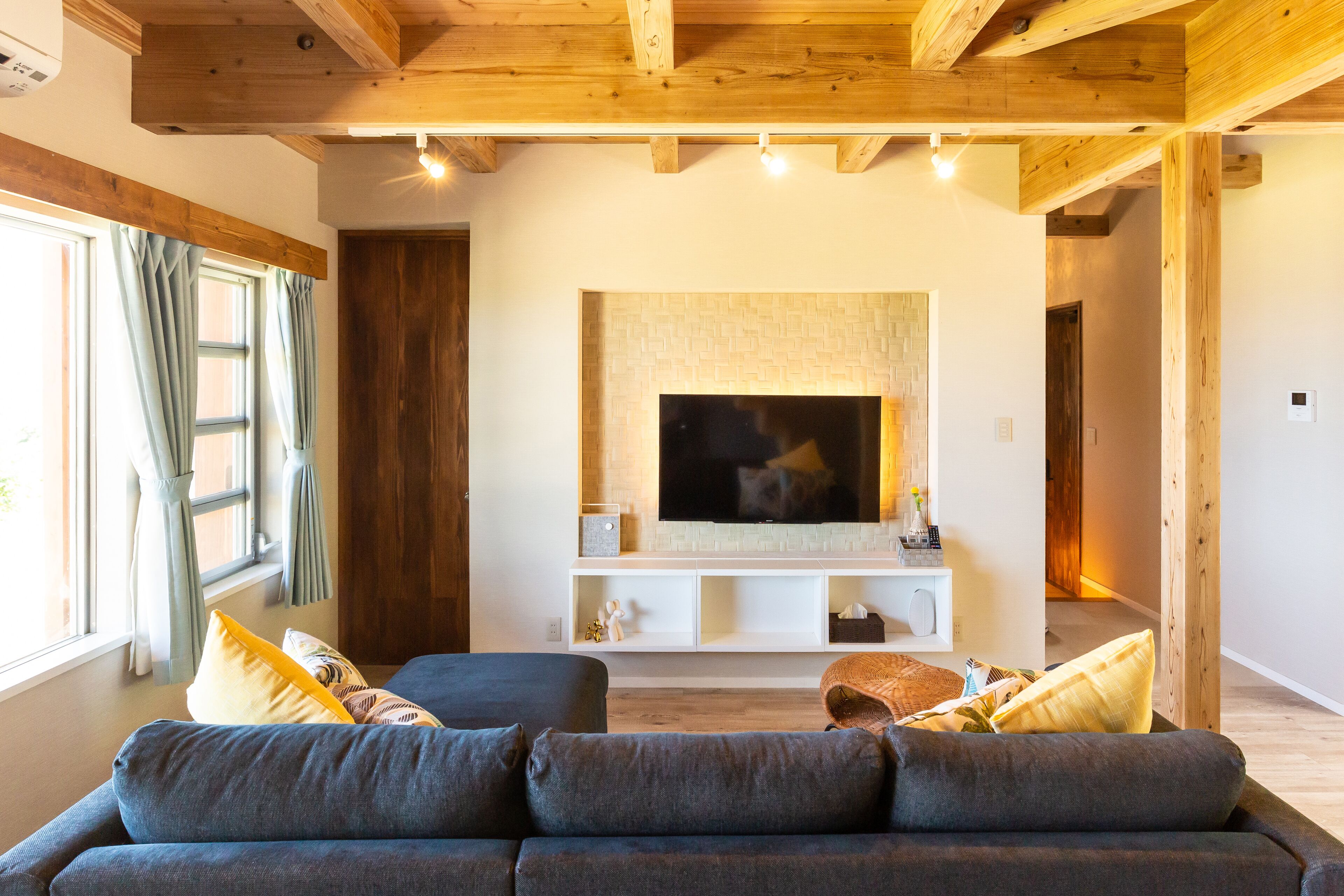 A modern living room with a large flat-screen TV, wooden ceiling beams, and a cozy seating area with a sofa and cushions.