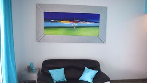 En svartvit soffa med två blå kuddar, ett litet sidobord med ett ljus och en inramad tavla som föreställer ett landskap med en båt på ett vattendrag.