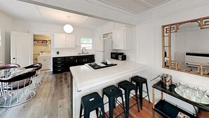 A modern kitchen with white cabinetry, a central island with stools, a dining area with a round table and chairs, and a large mirror with an ornate frame. - Charming Beach Cottage, 2 blocks from the water! (Morro Bay)