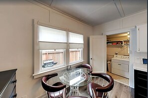 Eine kleine Küche mit Essbereich, ausgestattet mit einem Glastisch und vier Stühlen, einem weißen Herd und einem Fenster mit Jalousien.