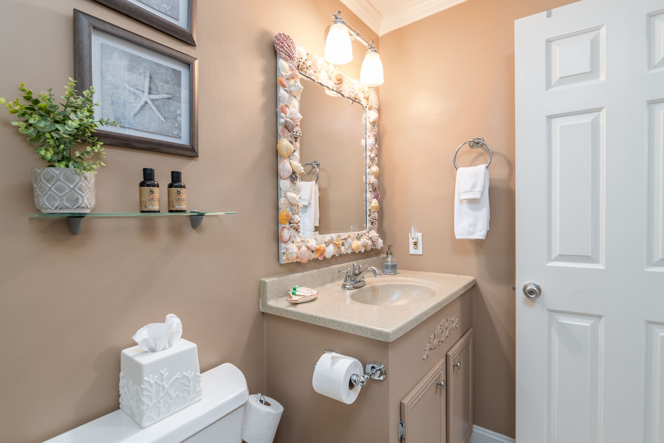 A bathroom with a beige wall, a white door, a sink with a mirror above it, and a shelf with toiletries.