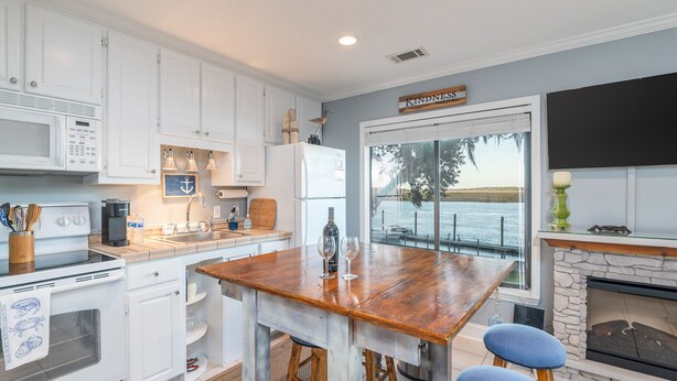 A modern kitchen with white cabinetry, a wooden island with stools, a stone fireplace, and a view of the ocean through large windows. - Gorgeous Waterfront Battery Creek Retreat (Beaufort)