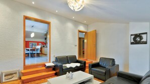Een moderne woonkamer met een bank, een salontafel, en een zichtbare eethoek door een grote spiegel.
