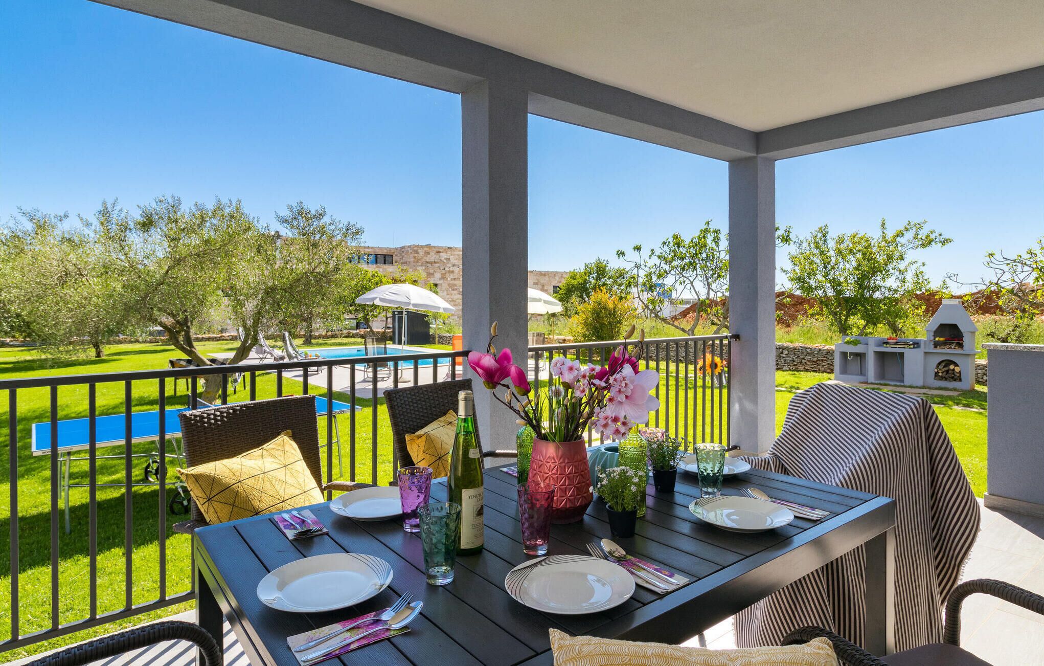An outdoor dining area with a table set for two, surrounded by chairs, a pool, and a barbecue grill.