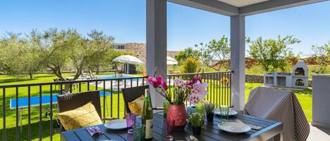 An outdoor dining area with a table set for two, surrounded by chairs, a pool, and a barbecue grill.