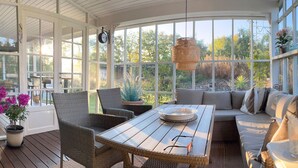 A sunroom with a wooden table, wicker chairs, a sofa, and large windows offering a view of trees and a garden.