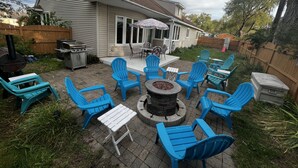 Un jardin avec un espace patio comprenant des chaises Adirondack bleues, un foyer en pierre et un barbecue.