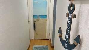 Un couloir doté d’un plancher en bois, d’un tapis turquoise, d’une porte ornée d’une fresque représentant une plage et d’une décoration en forme d’ancre portant l’inscription « BIENVENUE ».