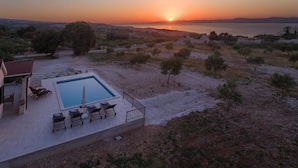 Un espace piscine doté de chaises longues et d’une table, entouré d’un paysage naturel avec des arbres et offrant une vue sur le coucher de soleil sur la mer.