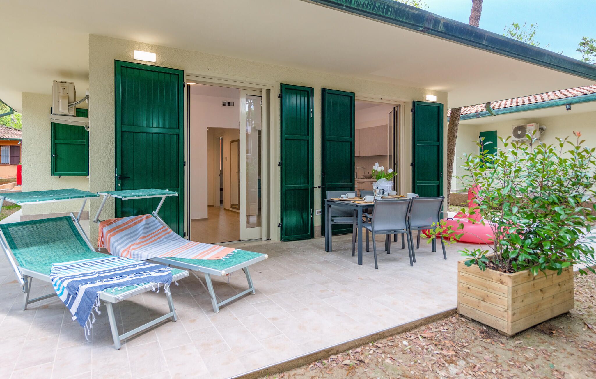 Un espace extérieur ensoleillé comprenant deux chaises longues, une table avec des chaises et un bac à fleurs en bois orné de feuillage verdoyant.