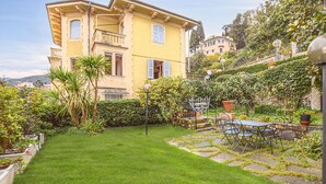 Un bâtiment à deux étages doté d’un balcon, entouré d’un jardin bien entretenu avec une pelouse et un chemin en pierre.