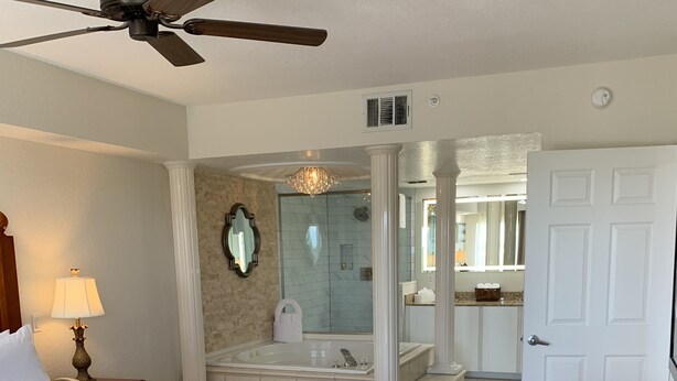 Une chambre d’hôtel avec un grand lit, une table en bois, un ventilateur de plafond, une salle de bain avec une baignoire et une douche.