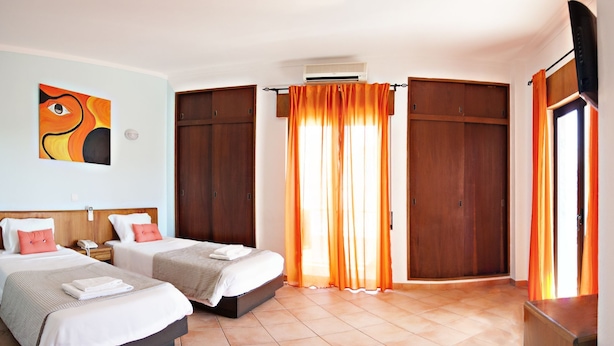 Ein Hotelzimmer mit zwei Betten, einem großen Fenster mit orangenen Vorhängen, einem Holzschrank und einem Gemälde an der Wand.