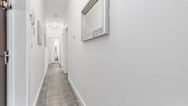 Un couloir étroit aux murs blancs et au sol carrelé, qui mène à une porte au fond.