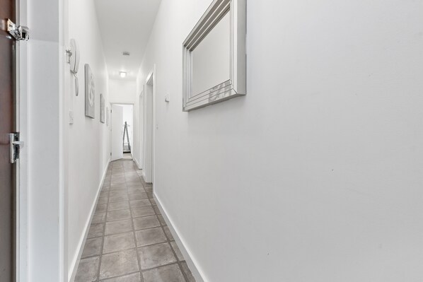 A narrow, white-walled hallway with a tiled floor, leading to a door at the end.