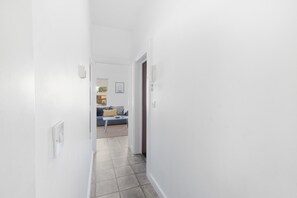 Un couloir étroit aux murs blancs et au sol carrelé mène à un salon où se trouvent un canapé bleu et une petite table.