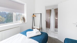 Une chambre avec un lit, une fenêtre avec un store, un radiateur, une porte donnant sur une salle de bain et un porte-serviettes avec des vêtements.