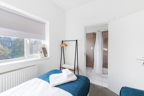 A bedroom with a bed, a window with a blind, a radiator, a door leading to a bathroom, and a towel rack with clothes.