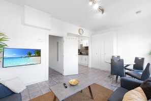 A modern living room with a flat-screen TV, a dining area with a glass table, and a kitchen in the background.